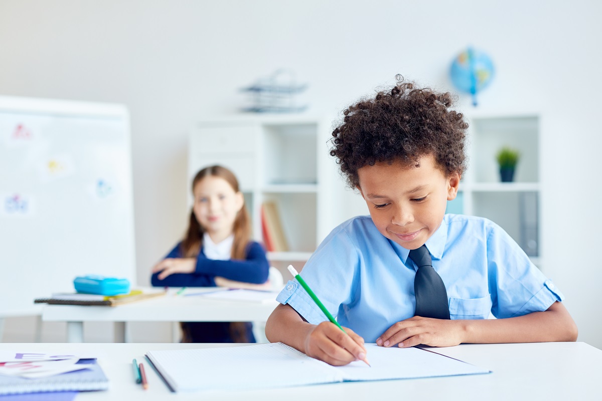 Little boy with pencil drawing or writing in notebook during lesson with schoolgirl on background